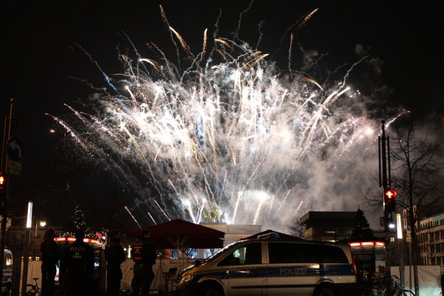Polizeiauto vor einer Menschenmenge während einer Silvesterfeier in Berlin geparkt, mit farbenfrohen Feuerwerken am Himmel.