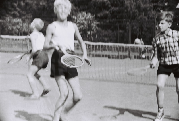 Drei Kinder spielen Tennis auf einem Platz, jedes hält einen Schläger in der Hand, mit einem Netz, einigen Menschen und Bäumen im Hintergrund.