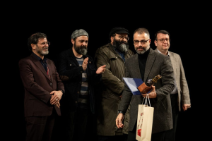 Eine Gruppe von Männern steht auf einer Bühne, einer hält eine Tasche und eine Trophäe, mit einem dunklen Hintergrund, der die Eröffnung der Berliner Filmfestspiele suggeriert.