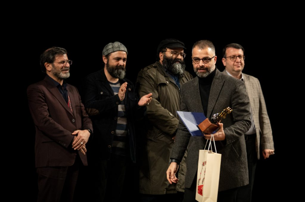 Eine Gruppe von Männern steht auf einer Bühne, einer hält eine Tasche und eine Trophäe, mit einem dunklen Hintergrund, der die Eröffnung der Berliner Filmfestspiele suggeriert.