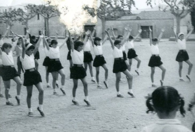 Schwarzes und weißes Foto von jungen Mädchen in Schuluniformen, die auf einer von Gebäuden und Bäumen gesäumten Straße tanzen, mit sichtbaren Personenschuhen unten.
