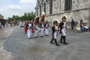 Eine Gruppe uniformierter Personen, einige im Rollstuhl und andere mit Gewehren, marschiert auf einer Straße in einer Militärparade in Frankreich vorbei an einem Gebäude, Bäumen und einem bewölkten Himmel.