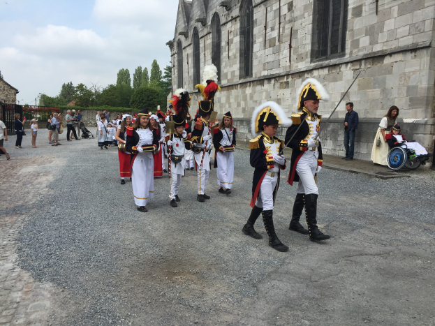 Eine Gruppe uniformierter Personen, einige im Rollstuhl und andere mit Gewehren, marschiert auf einer Straße in einer Militärparade in Frankreich vorbei an einem Gebäude, Bäumen und einem bewölkten Himmel.
