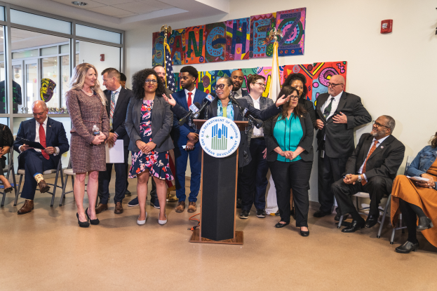 Eine Gruppe von Menschen, die in einem Raum mit einem Podium, einem Mikrofon und einer Tafel mit der Aufschrift "Sancho-Stadtrat tritt dem Stadtrat bei" stehen und sitzen, mit Flaggen, Feuerlöscher und Deckenleuchten an der Rückwand.