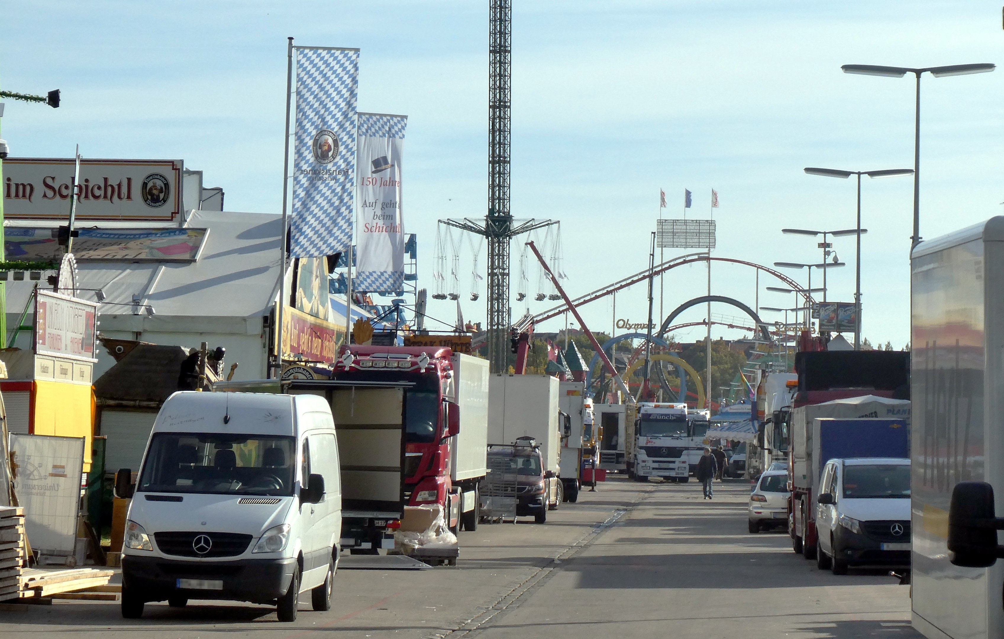 Vollgestopfte Straße mit Lastwagen, Autos, Fußgängern, Laternen, Bannern, einem Turm, einer Attraktion, Bäumen und einem bewölkten Himmel.