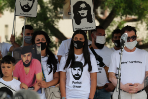 Eine Gruppe von Menschen mit Masken und Schildern vor einer Menge, mit Mikrofon und Stativ im Vordergrund, Bäume und Gebäude im Hintergrund und einer Kamera unten.