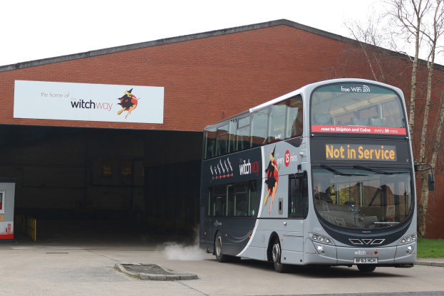Doppeldeckerbus mit Rauch auf einer Straße neben einem Gebäude fahrend, mit Gras, Bäumen und einem klaren blauen Himmel im Hintergrund und einem sichtbaren Fahrzeugkennzeichen am Bus.